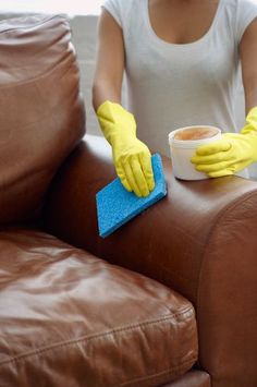 a woman in yellow gloves is cleaning a brown leather couch with a blue micro towel