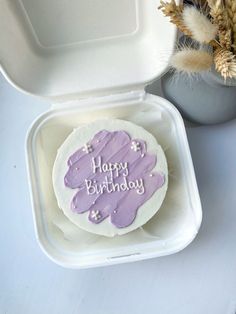 a purple frosted birthday cake sitting in a white container next to a flower pot