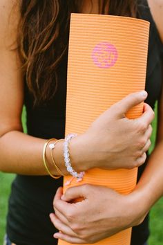 a woman holding an orange yoga mat in her right hand and bracelets on her left wrist