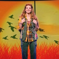 a woman standing in front of a screen with birds flying above her and an orange sunset behind her