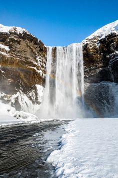 a large waterfall with a rainbow in the sky and snow on the ground next to it