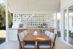 a wooden table sitting under a white pergoline covered outdoor kitchen with an oven in the background