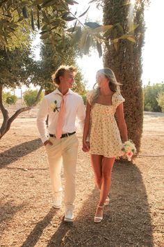 a man and woman are walking together in the dirt with trees on either side of them