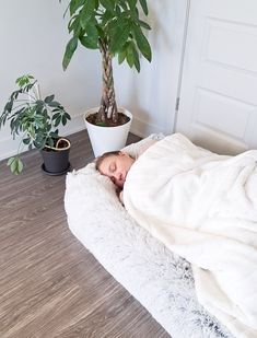 a woman laying in bed under a blanket next to a potted plant on the floor