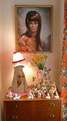 a wooden dresser topped with figurines next to a painting and vase filled with flowers