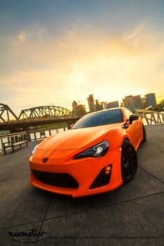 an orange sports car is parked in front of a bridge with the sun setting behind it