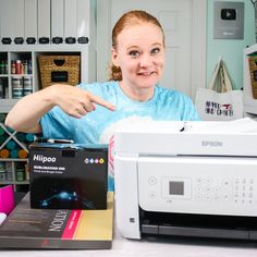 a woman pointing at a printer on the table next to it is another printer in front of her