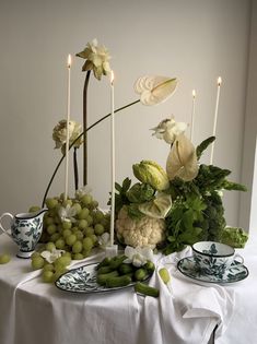 an arrangement of flowers, grapes and other food on a table with candles in the background