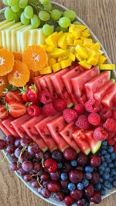 a platter filled with different types of fruit on top of a wooden table next to grapes, oranges, raspberries and watermelon