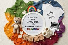 a baby announcement surrounded by clothes, toys and other items on a white surface with rainbow lights