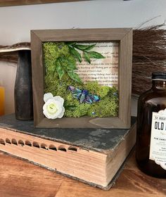 an old book with moss and flowers on it next to a bottle of orange juice