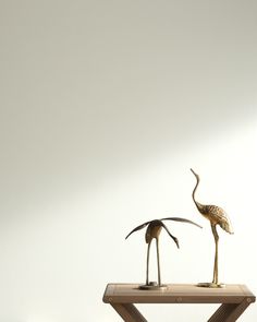 two metal birds standing on top of a wooden table in front of a white wall