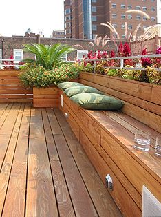 a wooden deck with planters and benches on the side, in front of tall buildings