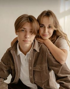 two young people sitting next to each other in front of a white wall and one is holding his arm around the other's shoulder