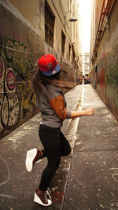 a young woman wearing a red hat and black pants is dancing in an alleyway