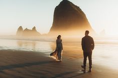 two people are standing on the beach looking at the water and mountains in the distance