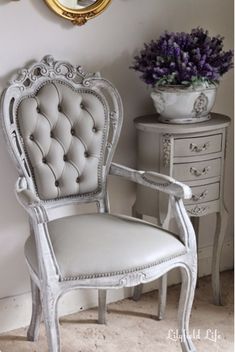 a white chair sitting in front of a mirror next to a vase with purple flowers