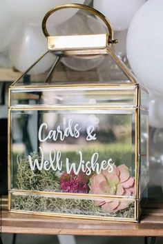 a clear glass box with cards and well wishes written on the front is surrounded by white balloons