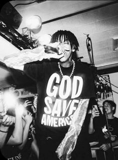 a young man standing in front of a crowd holding a microphone and wearing a t - shirt that says god save america