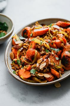 a bowl filled with carrots and nuts on top of a white countertop next to a plate of salad