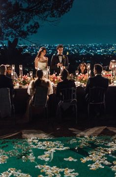 a group of people sitting around a table with flowers on the ground and lights in the background