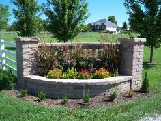 a brick wall with flowers in the middle and grass around it, near a white fence