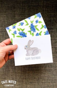 a hand holding up a card with a bunny on it and blue flowers in the background