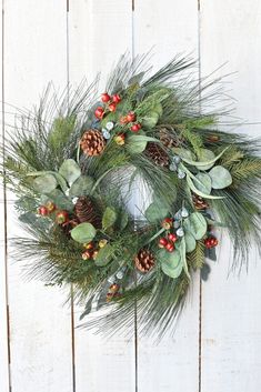 a green wreath with pine cones, berries and greenery hanging on a white fence