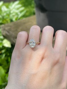 a woman's hand with a diamond ring on top of her finger, in front of some plants