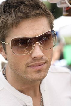 a close up of a person wearing sunglasses and a white shirt with people in the background