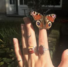 a woman's hand with two rings and a butterfly on the middle finger, in front of a house