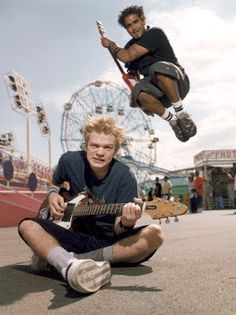 two young men are playing guitars in front of an amusement park