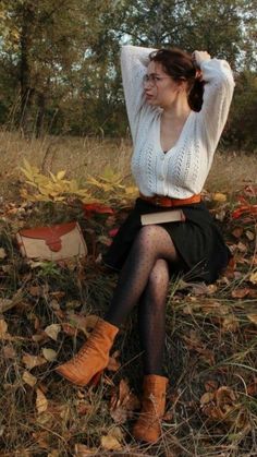 a woman sitting on top of a pile of leaves in the woods wearing boots and a sweater