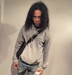 a young man with long hair standing in front of a white wall wearing jeans and a hoodie