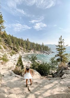 a girl in a white dress and hat walking up a hill near the water's edge