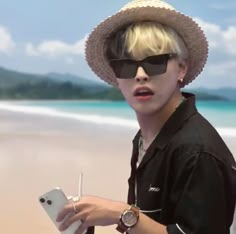 a man with sunglasses and a straw hat is holding a cell phone on the beach