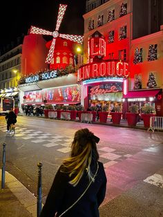 a woman standing on the side of a road next to a red building with christmas lights