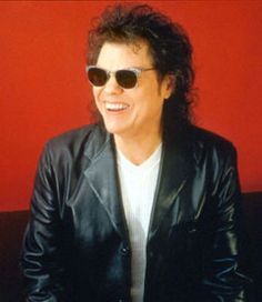 a man wearing sunglasses and a leather jacket sitting in front of a red wall smiling