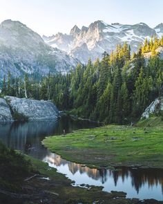 the mountains are covered in snow and green grass near a small lake surrounded by trees