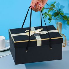 a woman's hand is holding a black box with a gold ribbon on it