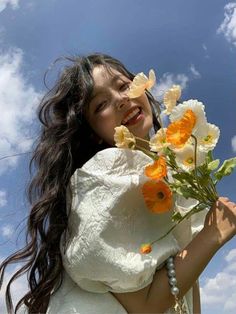 a woman holding flowers in her arms and smiling at the camera, against a blue sky with clouds