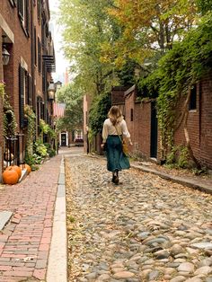 a woman walking down a cobblestone street next to brick buildings and pumpkins