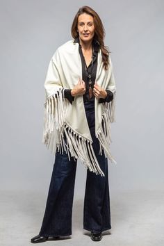 a woman standing in front of a gray background wearing a white shawl with fringes