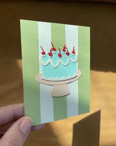 a hand holding up a card with a blue cake and cherries on the top