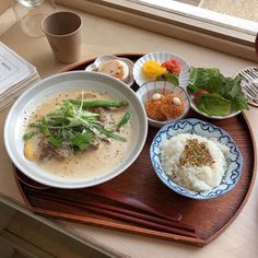 a wooden tray topped with bowls and plates filled with different types of food next to a window