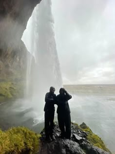 two people are standing in front of a waterfall and looking at the water from below