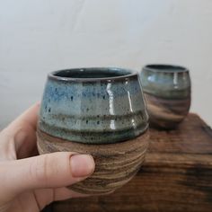 a hand holding a blue and brown cup on top of a wooden table next to another cup