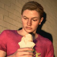 a young man is eating an ice cream cone in front of a brick wall and looking at his cell phone