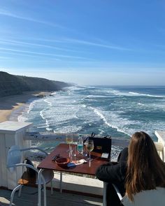 a woman sitting at a table on top of a wooden deck overlooking the ocean and beach