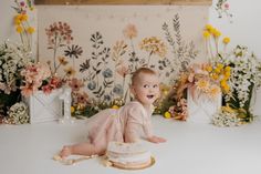 a baby is sitting on the floor next to a cake with flowers in the background